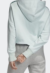 Krátký kapucový svetr v světle modré barvě, který má jemnou texturu, žebrované manžety a klokaní kapsu. Na zadní straně není viditelné logo.