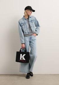 Lyseblå jeansjakke over en stripete skjorte, kombinert med vide jeansbukser. Svart totebag med hvit «K» og tegneserie-detalj. Svart caps.