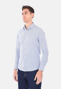 Camicia a maniche lunghe da uomo, azzurra chiara con righe verticali bianche, colletto alla francese, materiale in misto cotone e design aderente. Abbinata a jeans scuri.