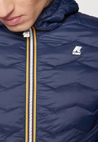 Veste rembourrée bleu marine avec une texture chevron, dotée d'une fermeture éclair à l'avant avec une rayure multicolore et d'un logo K-Way sur la poitrine.