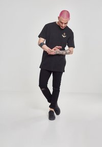Černé tričko s zlatým logem kotvy, oversize design, kombinováno s roztrhanými černými džíny a černými teniskami. Jednoduché pozadí.