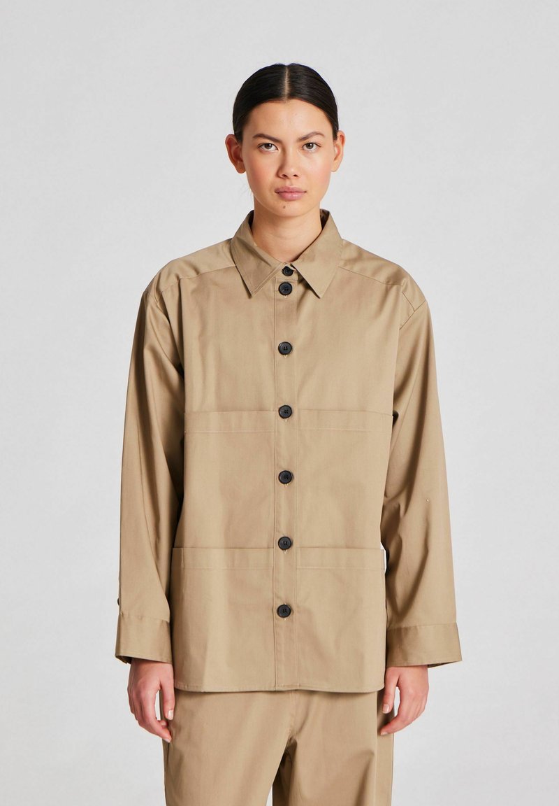 Tan button-up takki, jossa on kaulus, mustat napit, pitkät hihat ja sileä pinta. Minimalistinen muotoilu, jossa on kaksi vaakasaumaa.