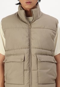 Beige puffer vest with high collar, front zipper, and two large button-flap pockets. Smooth texture with quilted padding.
