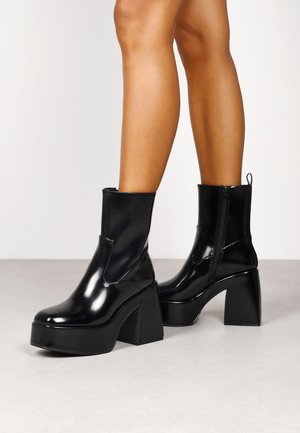 Botas de tobillo de charol negro con un alto tacón cuadrado, cremallera lateral y puntera redondeada, con un acabado elegante y brillante.
