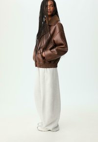 Donna con lunghe trecce che indossa una giacca di pelle marrone con colletto in shearling, pantaloni larghi grigio chiaro e sneakers bianche, in piedi di lato.