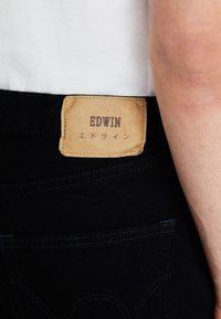 Tmavě modré džíny z denimu s hrubou koženou nášivkou v barvě tan, na které je vytištěno "EDWIN" a japonský text; mají decentní švy a design s pěti kapsami.