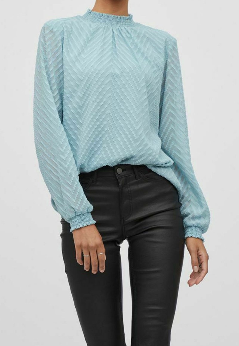 Femme portant un chemisier bleu clair texturé à manches longues, rentré dans un pantalon en cuir noir taille haute et coupe étroite.