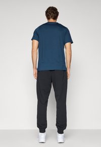 T-shirt med korte ærmer i mørk teal, parret med løstsiddende sorte joggingbukser, båret med hvide sportssko. Minimalistisk design, glat tekstur.