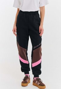 Osoba nosząca czarne dresy z brązowymi i różowymi ukośnymi blokami kolorów oraz tekstem, białą koszulkę, różowe skarpetki i brązowe platformowe trampki.