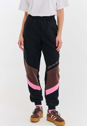 Osoba nosząca czarne dresy z brązowymi i różowymi ukośnymi blokami kolorów oraz tekstem, białą koszulkę, różowe skarpetki i brązowe platformowe trampki.