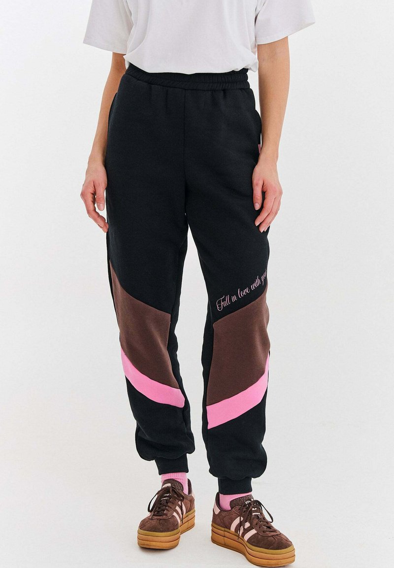 Osoba nosząca czarne dresy z brązowymi i różowymi ukośnymi blokami kolorów oraz tekstem, białą koszulkę, różowe skarpetki i brązowe platformowe trampki.