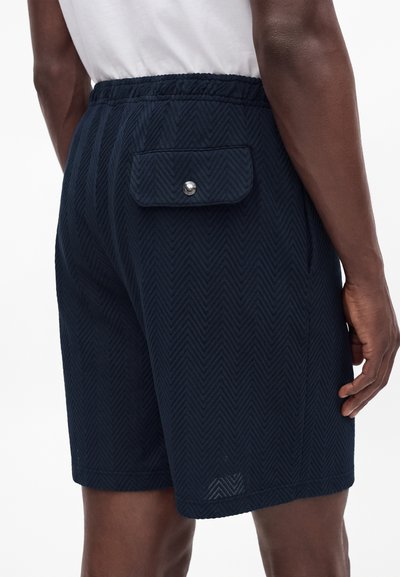 Homme portant un short bleu foncé texturé avec une taille élastique et une poche à rabat boutonnée à l'arrière, porté avec une chemise blanche.