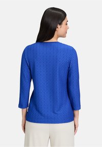 Blaues Langarmtop aus strukturiertem, gemustertem Stoff mit rundem Ausschnitt und geradem Schnitt. Sichtbare Nähte und Dreiviertelärmel.