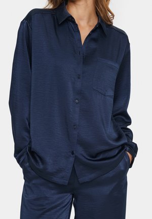 Kobieta ubrana w granatową jedwabną koszulę na guziki z kieszenią na piersi oraz dopasowane spodnie, z rękami w kieszeniach, na jednolitym tle.