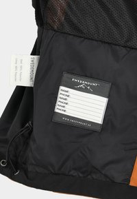 Black and orange jacket featuring a mesh lining, fabric tags, and a label for personal details with a mountain logo.