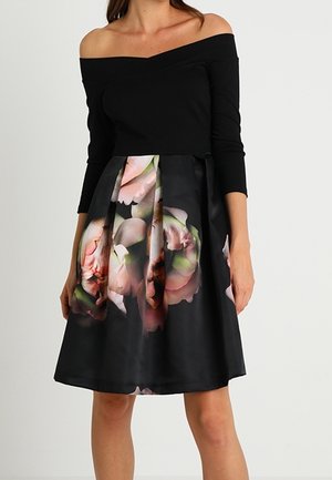 Femme portant une robe noire épaules dénudées avec des manches trois-quarts et un motif floral sur une jupe plissée, debout devant un fond uni.