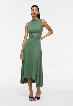Femme portant une robe midi asymétrique verte sans manches et des talons noirs ouverts, posant devant un fond uni clair.