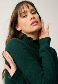 Maglione a collo alto verde scuro con una texture morbida e scollatura a onda. Le maniche lunghe presentano polsini a costine. Design semplice e aderente.