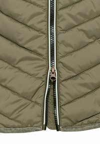 Olivgrüne gesteppte Jacke mit einem Frontreißverschluss und bronzenen Beschlägen, kontrastierenden schwarzen Zähnen und strukturiertem Stoff mit Zickzackmuster.
