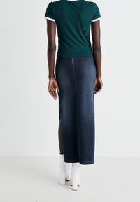 Camiseta verde ajustada con ribete blanco, combinada con una falda de mezclilla larga con una abertura lateral y llevada con botas de tobillo de tacón cuadrado blancas.