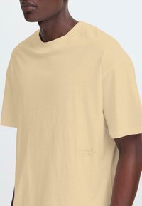 Licht beige t-shirt met korte mouwen van zachte katoen. Heeft een ronde halslijn en een subtiele geborduurde logo aan de zijkant. Soepele textuur.