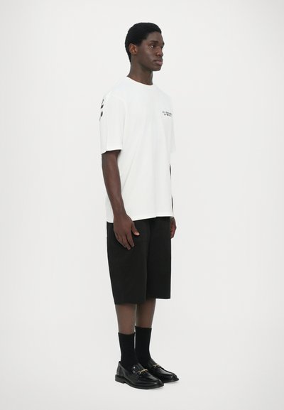 T-shirt blanc à manches courtes avec un texte graphique noir, associé à un short noir arrivant aux genoux et des mocassins noirs avec un accent doré.