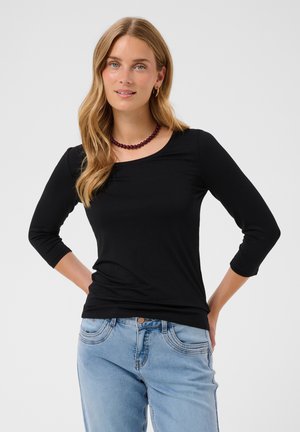 Vrouw met lang blond haar, die een zwarte top met driekwart mouwen, lichtblauwe spijkerbroek en een donkere kralenketting draagt, staand met handen in de zij.