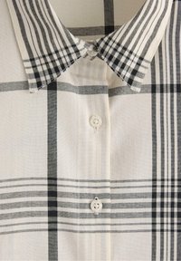 Vit skjorta med svart och grå rutmönster, med knappad krage och två synliga vita knappar längst fram.