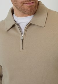 Béžový pulovr na čtvrt zip s límečkem, vyrobený z měkké tkaniny. Obsahuje stříbrný zip a hladký povrch. Přes bílou košili.