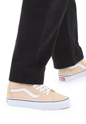 Zapatillas Vans beige con cordones blancos y franja lateral, llevadas con pantalones negros sobre un fondo blanco.