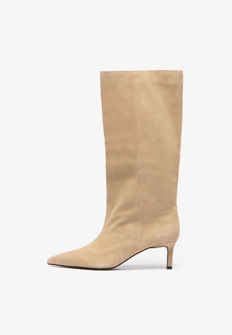 Botte en daim beige, montant jusqu'au genou, avec un bout pointu et un talon fin. Présente des coutures verticales le long de la tige pour un détail supplémentaire.