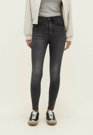 Personne portant un jean skinny noir délavé taille haute avec des ourlets effilochés, un haut côtelé gris clair, une veste bomber crème et des baskets noires et blanches.