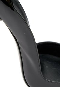 Gros plan d’une chaussure à talon haut en cuir noir montrant un talon courbé lisse et des bords cousus sur fond blanc.