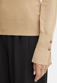 Maglione lavorato a maglia beige con orlo e polsini a coste, dotato di due bottoni marroni sul maniche sinistro. Il marchio "Guess" è ricamato in modo discreto.