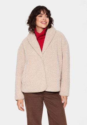 Beige teddy jas van een gestructureerde, donzige stof. Beschikt over een sjaalkraag, open voorkant en twee zijzakken. Draag het over een rode coltrui.