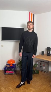 Svart långärmad tröja med stort tryck av "XOXO", kombinerad med mörka jeans med vida ben och bruna läderloafers. Orange lampa i bakgrunden.