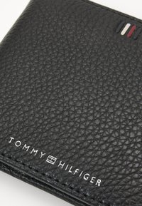 Portafoglio in pelle nera con una texture a ciottoli, bordato da una cucitura. Accentuato da piccole strisce rosse, bianche e blu e dettagli con logo.