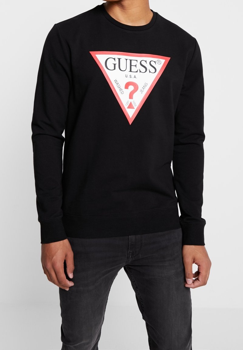 Man in een zwart sweatshirt met een groot rood en wit driehoekig Guess-logo en vervaagde zwarte jeans tegen een effen achtergrond.