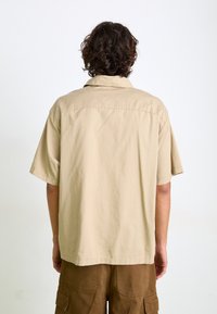 Short-sleeve, light tan shirt with a back yoke and a pointed collar, made from smooth fabric. Worn with brown cargo pants, showing seam details.