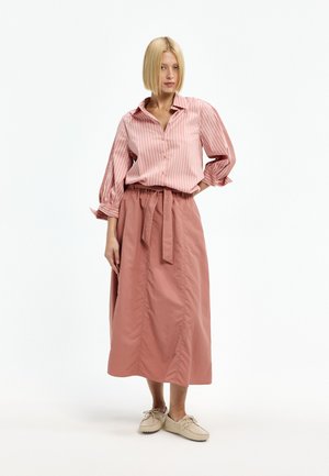 Frau mit blondem Bob, die eine rosa gestreifte Bluse, einen hoch taillierten, gebundenen Midirock und beige Schnürschuhe trägt, steht mit der Hand in der Hüfte.