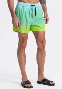 Costumi da bagno in gradiente turchese verso verde, con vita elasticizzata, cordino regolabile e texture liscia. Includono sandali neri.