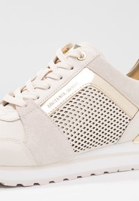 Baskets beige clair combinant des matériaux lisses et en suède, avec une plaquette décorative dorée, un panneau latéral perforé et une semelle texturée.