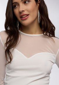 Top bianco a maniche lunghe con scollatura in rete trasparente e design a zigzag. Tessuto morbido con silhouette aderente. Orecchini scintillanti.