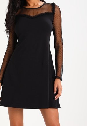 Vrouw draagt een zwarte jurk met doorschijnende, met stippen versierde lange mouwen en een doorschijnende inzette met gerafelde rand over de borst.