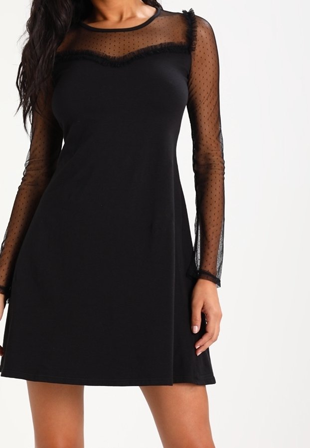 Femme portant une robe noire avec des manches longues en tulle transparent à pois et un empiècement transparent orné d'une bordure à volants sur la poitrine.