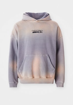 Hettegenser i lilla og fersken gradient, laget av myk stoff. Har en lommepung foran og logodetaljer i svart på brystet.