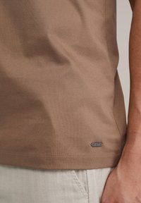 T-shirt in cotone marrone con una texture liscia e un piccolo tag metallico sul bordo. Design leggermente aderente con maniche corte.