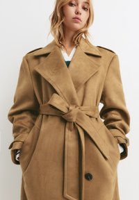 Manteau beige en daim doux, doté d'une ceinture, de grands revers, de poches latérales et d'un détail de bouton noir.