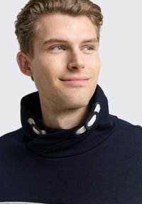 Navy Pullover mit hohem Kragen und cremefarbenen Akzenten. Verfügt über einen strukturierten Strickstoff und ein einzigartiges Zubehördetail in Form von Schlaufen am Kragen.