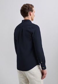 Chemise bleu marine à manches longues avec col boutonné, pli dos et poignets. Texture de tissu lisse, conçue avec une coupe ajustée.